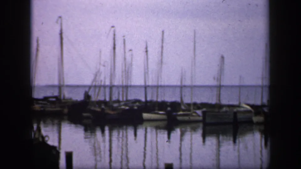 1949 Ireland 8mm frame showing docked boats, closely spaced masts, reflections, and a low shoreline beyond the harbor.