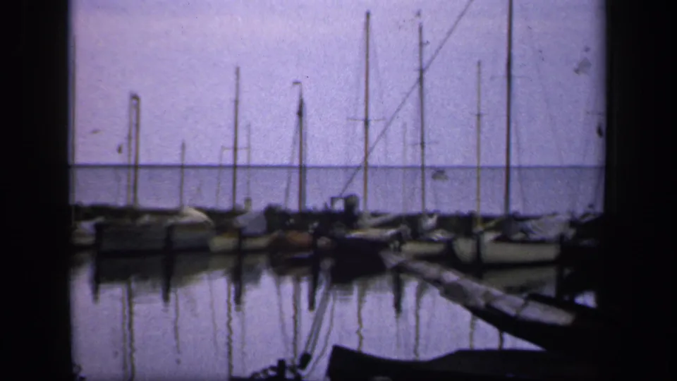 1949 Ireland home-movie frame looking past a wooden pier toward docked sailboats and reflected masts.
