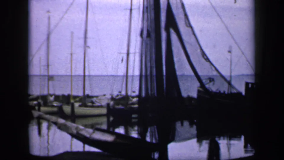 1949 Ireland 8mm frame showing dark fishing boats with rigging and nets at a calm harbor, with water reflections and small birds overhead.