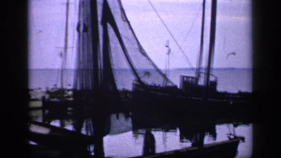 1949 Ireland harbor view with a silhouetted fishing vessel, high rigging, docked boats, calm reflections, and birds crossing the pale sky.