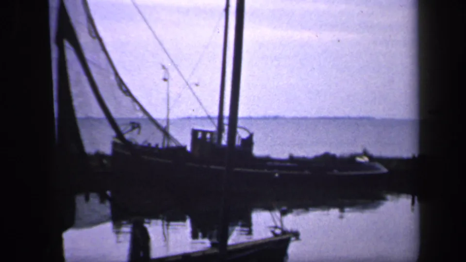 1949 Ireland 8mm frame showing a moored fishing boat with mast, rigging, and netting beside reflective harbor water and a low distant shoreline.