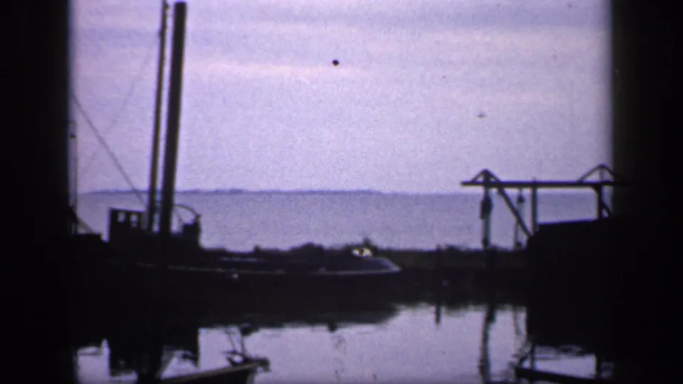 1949 Ireland harbor frame with a dark moored boat, vertical mast, simple dock structure, still water reflections, and a pale sky over distant land.