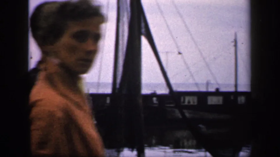 1949 Ireland home-movie frame with an adult face briefly visible near moored boats, rigging, and calm dockside water.