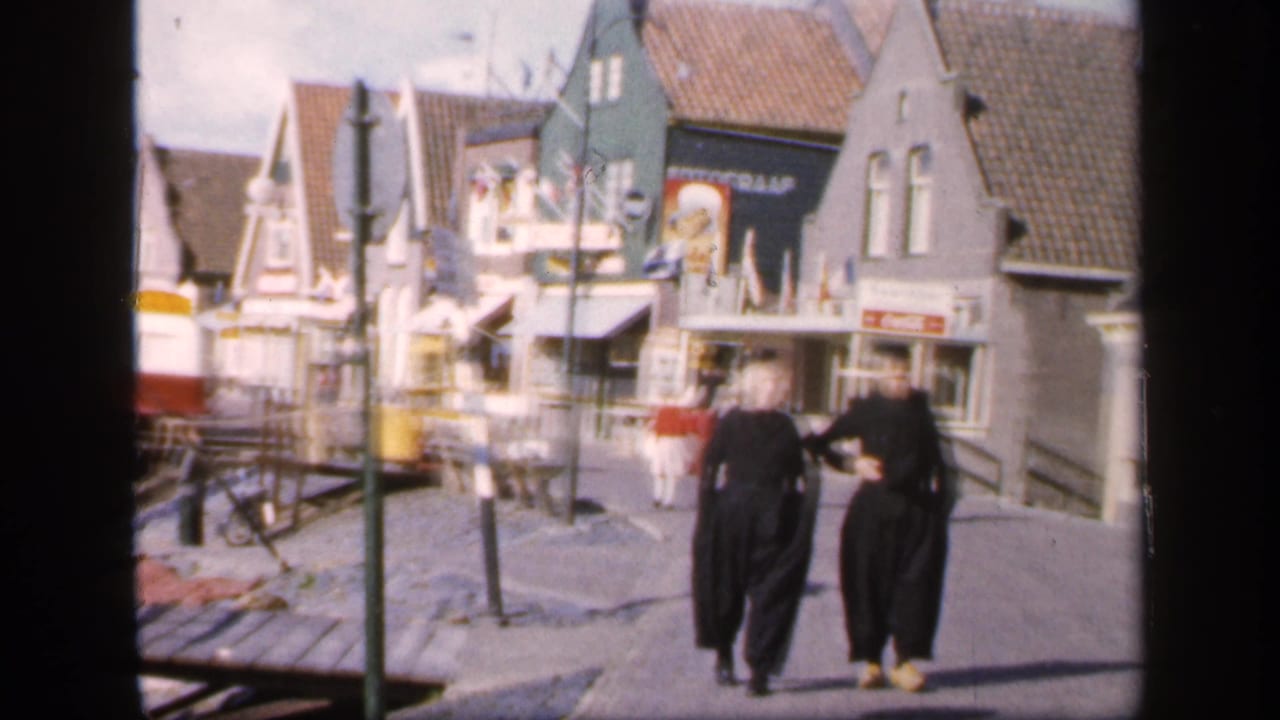 Two women walk along a cobbled waterfront street lined with gabled buildings and shopfronts.