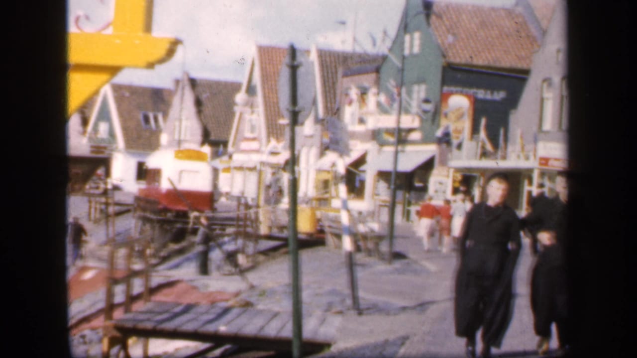Pedestrians walk near a small rail vehicle and gabled waterfront buildings.