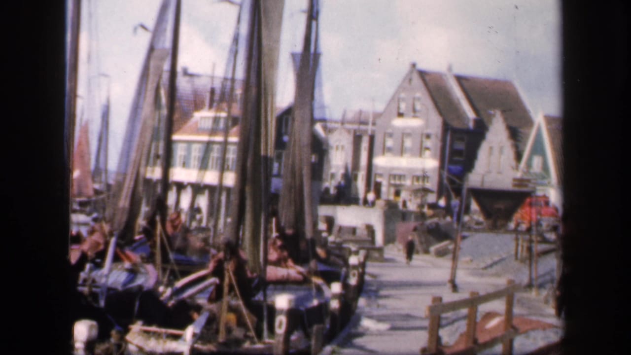 Quayside harbor view with sailboat masts, buildings, and people walking near the water.