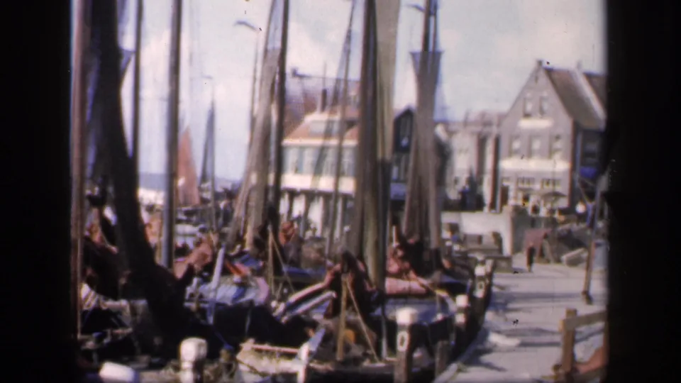 1949 Ireland 8mm frame from among moored boats, with rigging, a quay, distant people, and waterfront buildings visible.