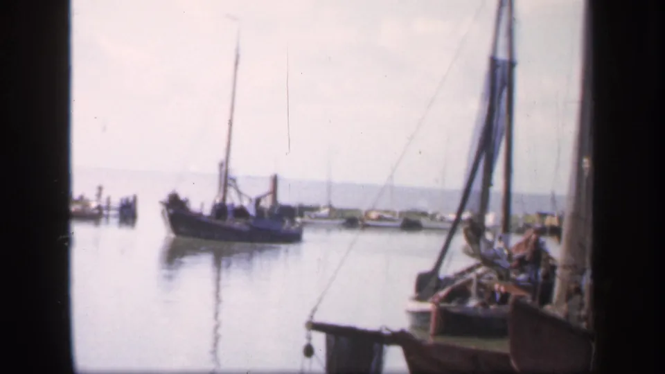 1949 Ireland 8mm frame of a wooden sailboat on smooth harbor water, with docked boats and a pale shoreline in the distance.