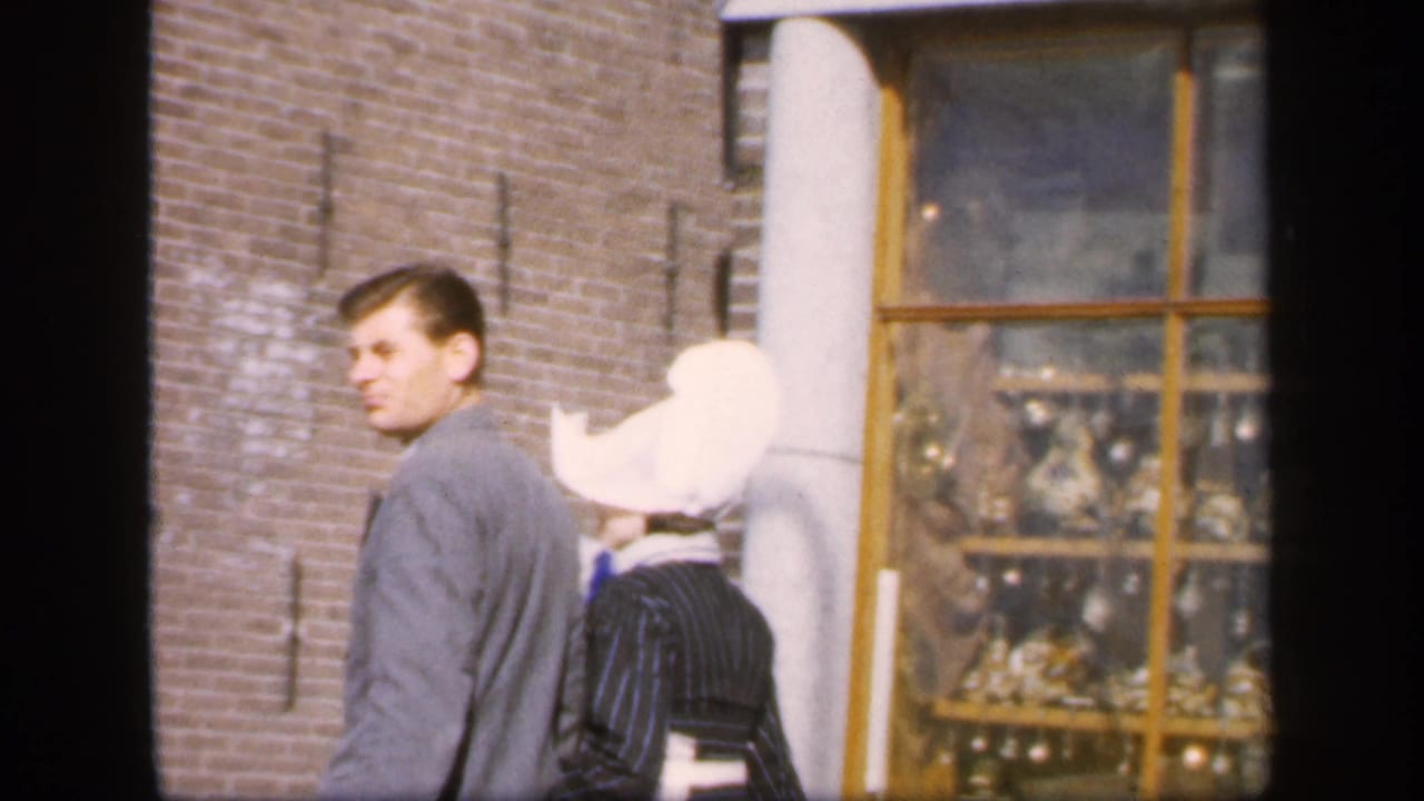 Man in a grey suit walks beside a woman in a white bonnet near a brick wall and shop window.