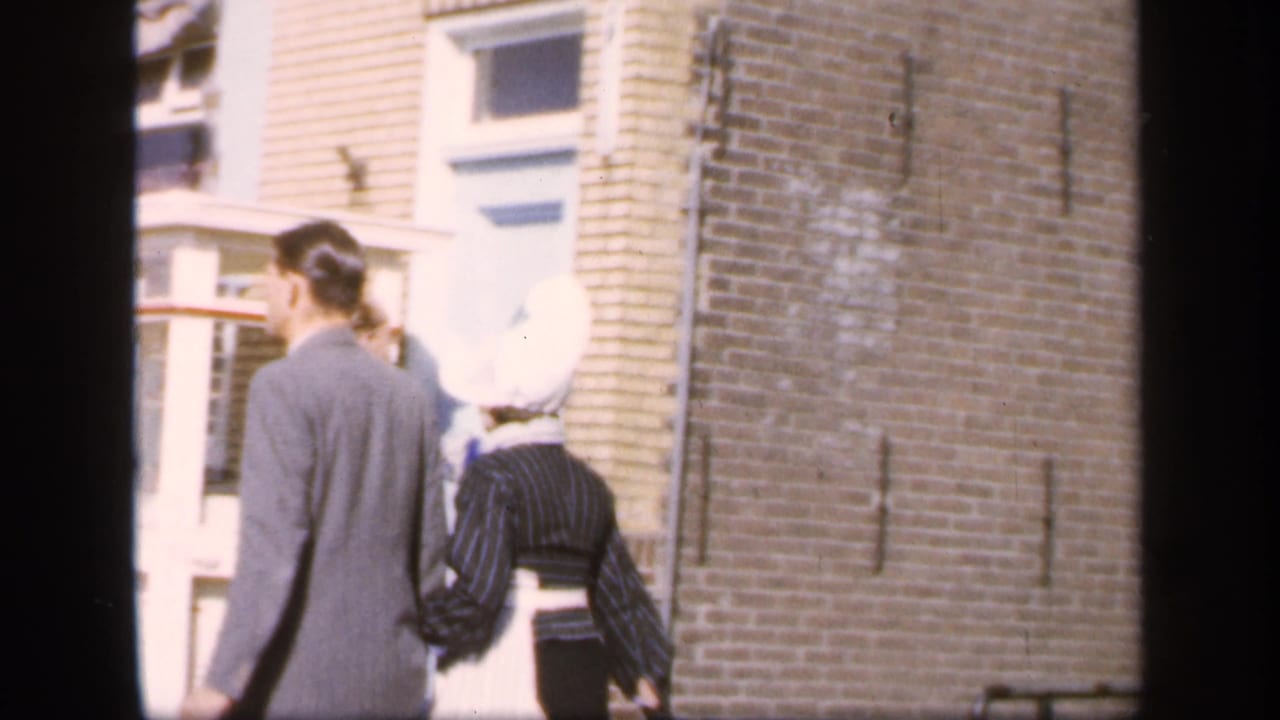 The pair walks away along a brick-lined street, with the woman’s white bonnet and striped dress visible beside the suited man.