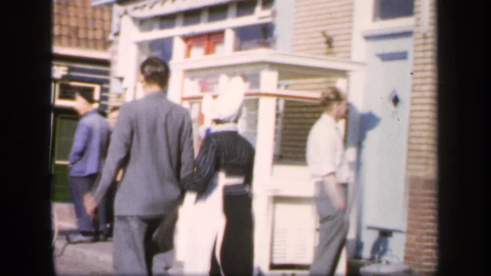 A small group of pedestrians is visible near a brick streetfront, with the bonneted woman and suited man continuing away from view.