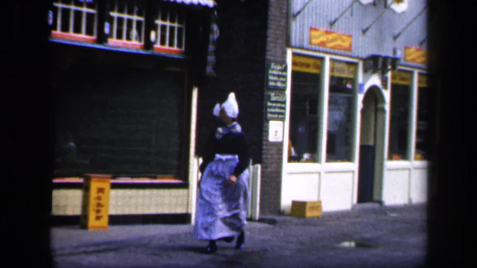 A bonneted woman in traditional-style clothing walks along a shaded street with storefront windows and hanging signs nearby.