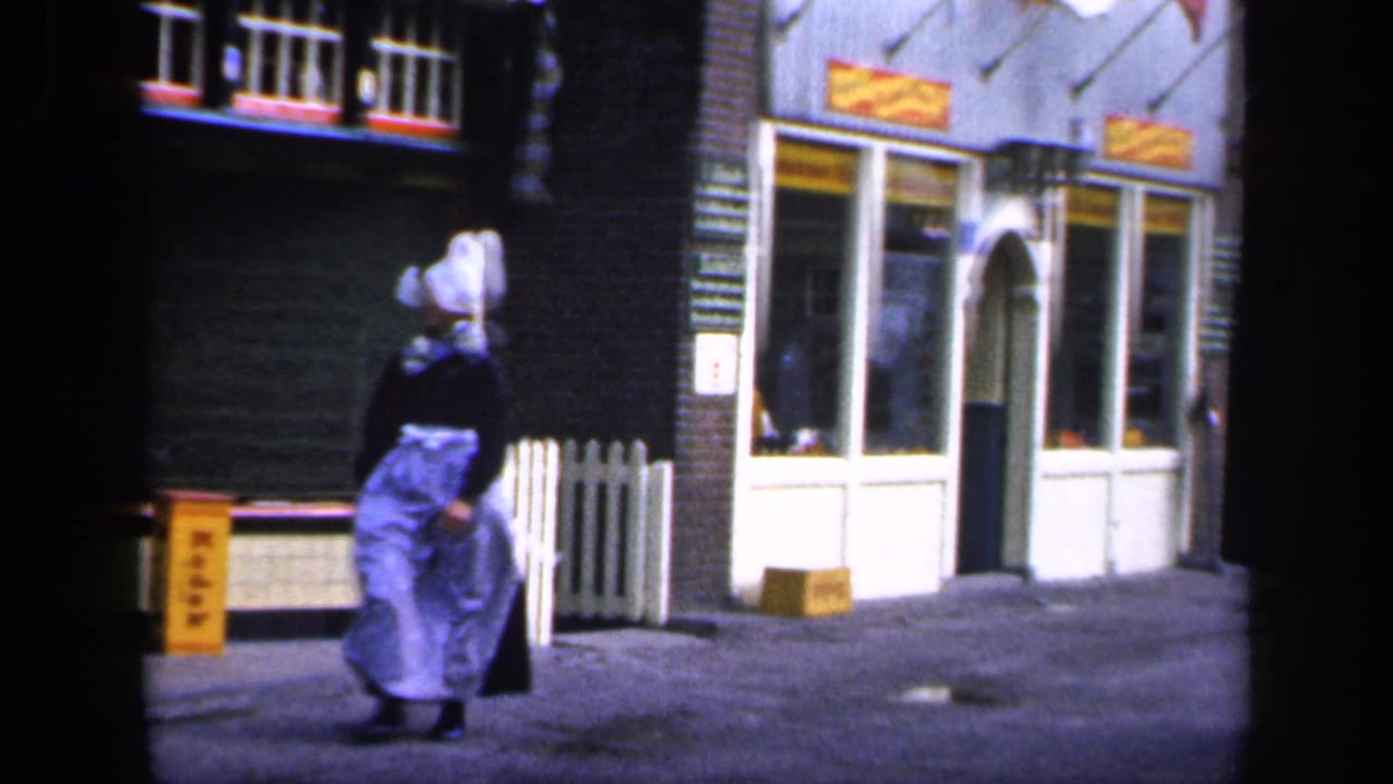 In Ireland archival 8mm footage, a woman in traditional dress walks past storefront windows on a quiet cobblestone street.