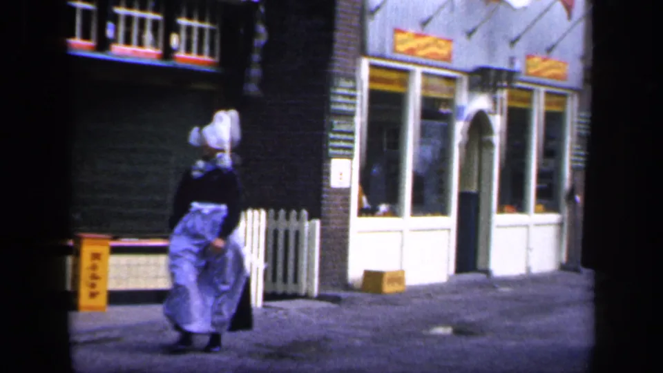 In Ireland archival 8mm footage, a woman in traditional dress walks past storefront windows on a quiet cobblestone street.