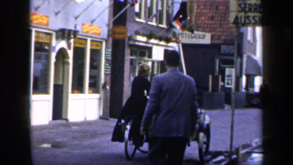 A man follows a woman with a bicycle sidecar past storefronts and signs in a 1949 Ireland street scene.