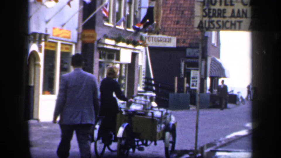 A man and a woman with a bicycle sidecar move down a narrow Irish street with shop signs and distant pedestrians.