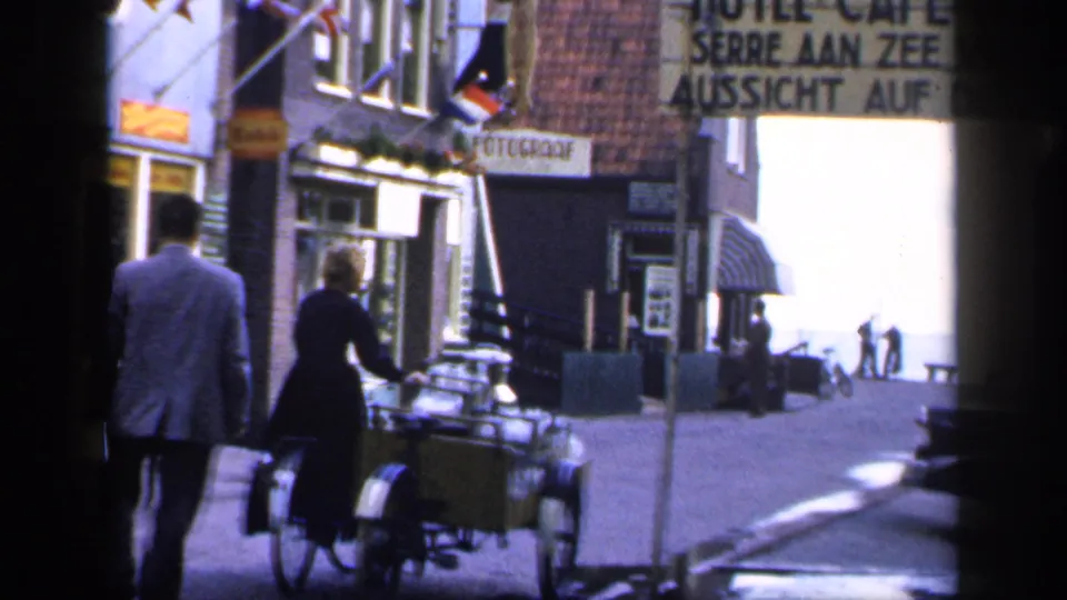 1949 home-movie frame showing pedestrians near a bicycle with cargo basket on a narrow European street with shop signs and buildings.