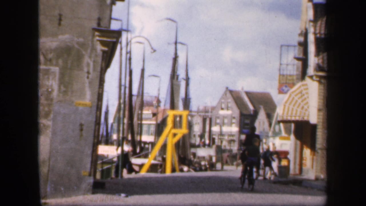 A narrow waterfront street with cyclists, pedestrians, buildings, and a yellow harbor-side structure.