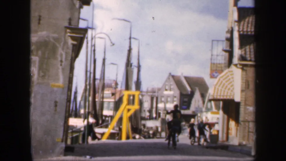 1949 home-movie frame showing a cyclist, pedestrians, harbor masts, and a yellow structure along a compact European street.