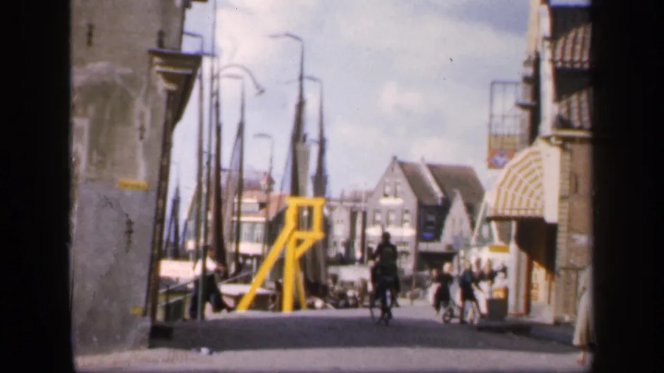 1949 home-movie frame of pedestrians and a cyclist on a European waterfront street with buildings, masts, and a yellow structure.