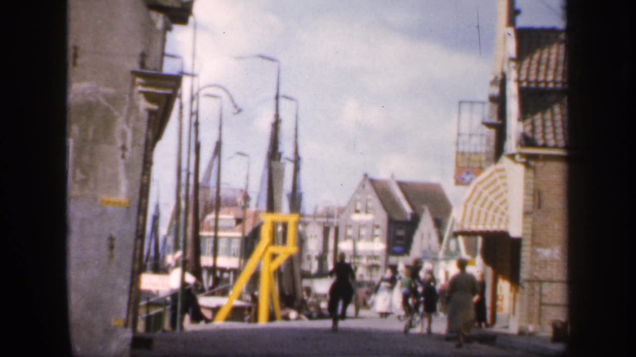 1949 Ireland home-movie frame showing pedestrians on a narrow town street with brick buildings, curved lamps, and a yellow bridge structure.