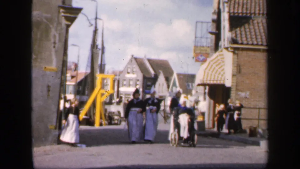 1949 Ireland street view with women in white aprons and bonnets walking toward the camera beside a cyclist and town buildings.