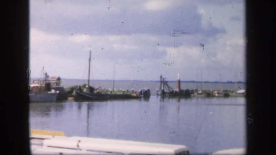 1949 Ireland home-movie frame with small boats and dock structures on calm harbor water beneath clouds.