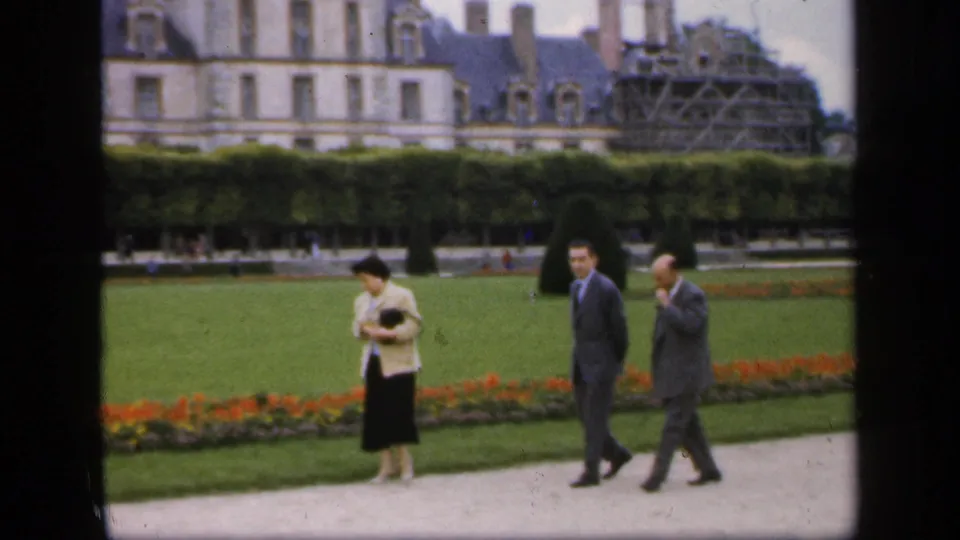 1949 Ireland home-movie frame showing adults strolling past formal gardens, clipped hedges, and a grand building in the background.