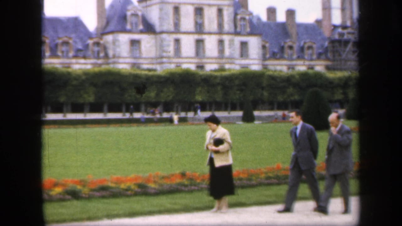 Woman and two men walking beside a flowerbed in front of a large château-like building.
