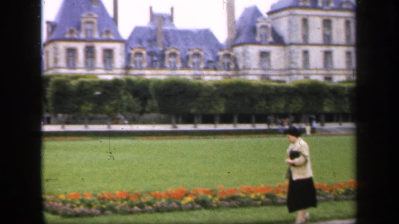 Woman walking alone along a formal garden path with lawns, flowers, hedges, and a large building behind.