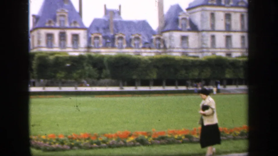 A woman in a light jacket and dark skirt walks through a formal garden, with a grand slate-roofed building in the background.