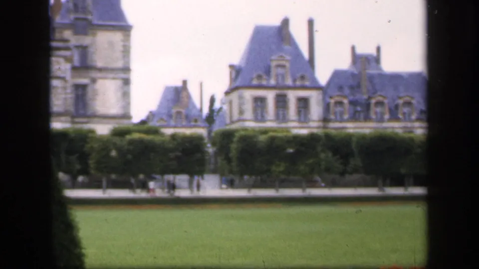 The camera frames the formal grounds and upper château-like facade, with visitors faintly visible near the hedge line.