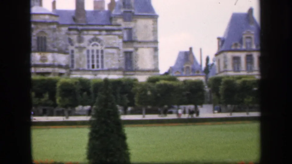 A formal garden view shows a conical topiary, clipped hedges, lawn, and a grand historic building beyond.