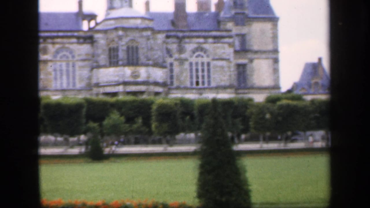 Grand stone chateau seen beyond lawns, hedges, flowerbeds, and dark window framing.
