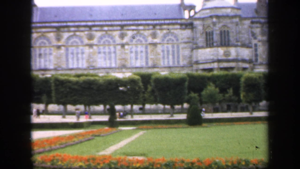 Long chateau facade, clipped hedges, lawn, flowerbed, paths, and distant visitors.