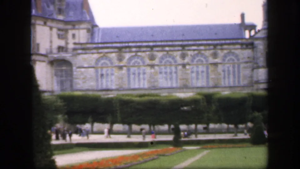 A soft 8mm frame shows the chateau wall above formal hedges and paths, with small groups of visitors crossing the garden area.
