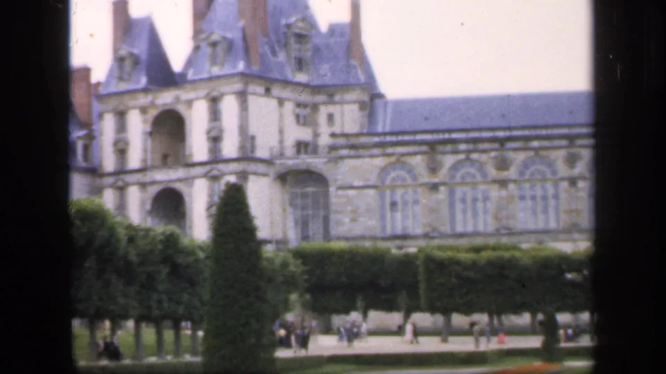 The final frame centers more of the ornate entrance and roofline, with clipped hedges, shaped trees, and distant visitors in the formal garden.