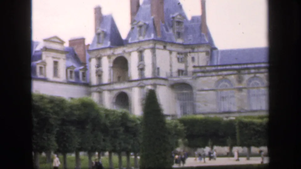 1949 8mm frame of a grand chateau-like building behind formal hedges, with visitors walking through the garden grounds.