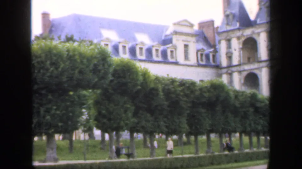 Clipped hedges fill the foreground as part of the chateau-like facade remains visible, with visitors moving along the garden path.