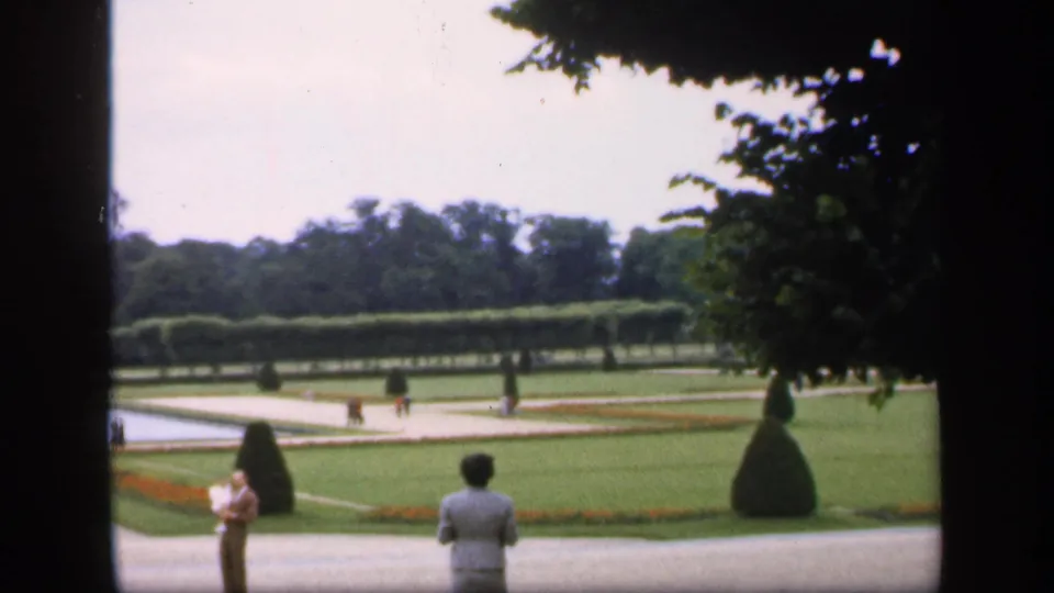 The view opens to formal lawns, geometric paths, trimmed hedges, topiary, and visitors standing or walking in the garden.