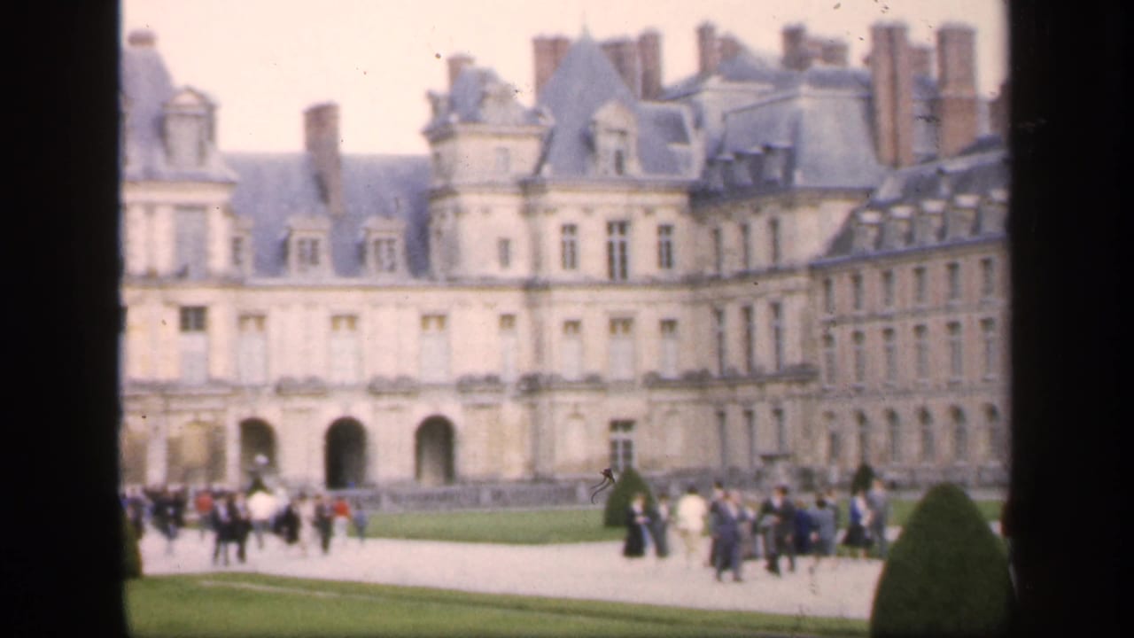 Grand stone castle facade with visitors walking across lawns and gravel paths.