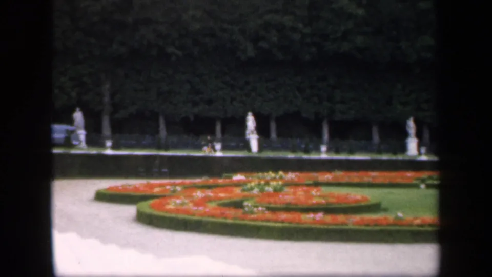 1949 8mm frame of a formal garden in Ireland, showing geometric red flowerbeds, low hedges, white statues, and a dense tree line.