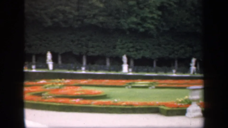 1949 Ireland home-movie frame showing formal red and orange flowerbeds, trimmed hedges, white statues, trees, and a partial stone garden feature.