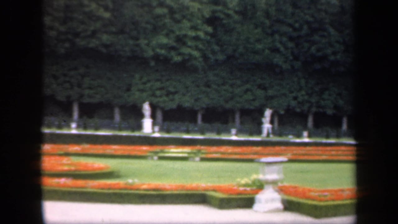 1949 Ireland 8mm frame showing a formal garden with orange flowerbeds, trimmed hedges, green lawn, distant statues, and a central pedestal.