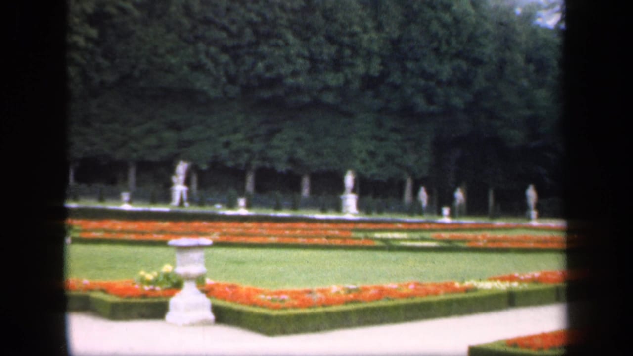 1949 Ireland garden view with geometric flowerbeds, clipped hedge borders, distant statues, pale walkways, and dark 8mm frame edges.