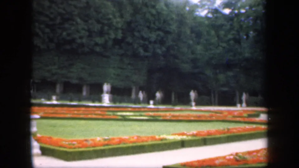 Soft 8mm frame from 1949 Ireland showing a formal garden, broad lawn, orange planting beds, clipped hedges, and small distant figures or statues.