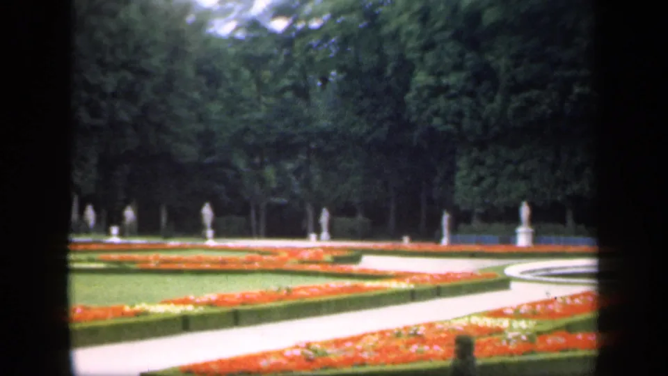 1949 Ireland 8mm view of a formal garden with curving pale path, orange flowerbeds, clipped hedge edges, dense trees, and distant statues.