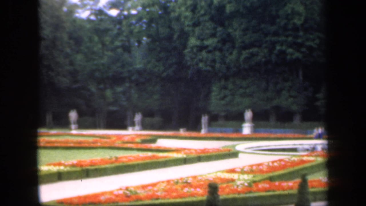 Formal garden with orange flowerbeds, pale paths, statues, lawns, and dense trees beyond a dark foreground frame.