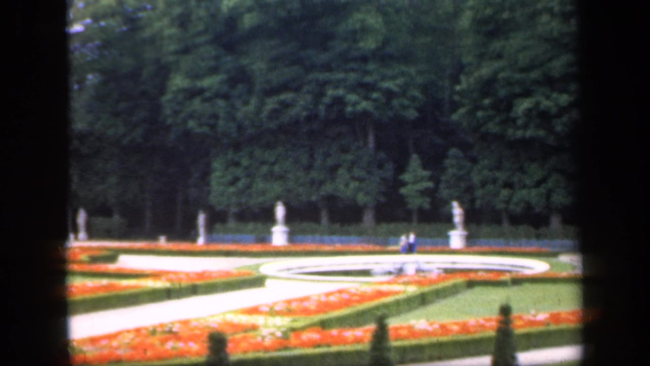 Formal garden with circular fountain area, orange flowerbeds, white statues, trees, and small distant visitors.