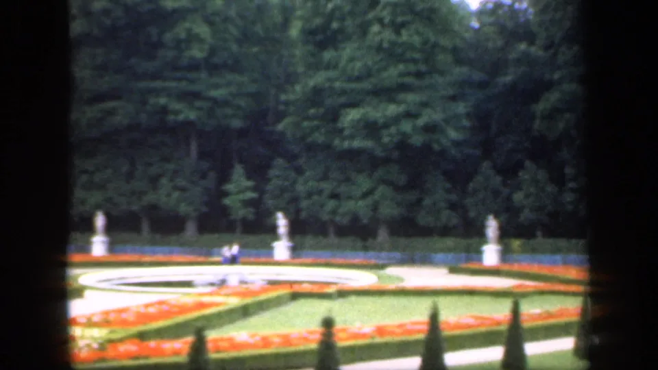 1949 Ireland 8mm frame of curved garden paths around a circular feature, orange flowerbeds, statues, trees, and distant visitors.